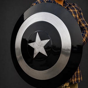 Puede incluir: Un escudo negro y plateado con un dise&ntilde;o de estrella plateada en el centro. El escudo tiene forma circular con un anillo plateado alrededor del borde. Es un accesorio o art&iacute;culo de colecci&oacute;n.