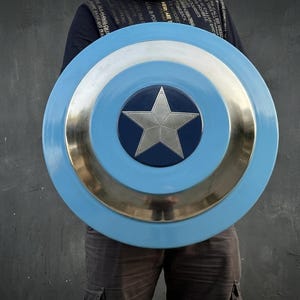 May include: A circular shield in light blue, silver, and dark blue. The shield has a silver star at its centre, encircled by silver and light blue rings. The shield is held in front of a person.