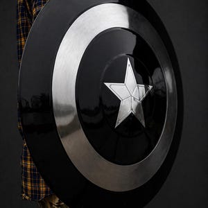 Puede incluir: Un escudo negro y plateado con un dise&ntilde;o de estrella central. El escudo tiene forma circular con un anillo plateado alrededor del borde. La estrella es plateada y tiene cinco puntas. Es un accesorio o art&iacute;culo de colecci&oacute;n.