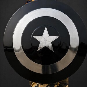 Puede incluir: Un escudo redondo negro y plateado, con una estrella plateada central. El escudo tiene un centro negro brillante, un anillo plateado y un borde exterior plateado. El escudo parece ser de metal.