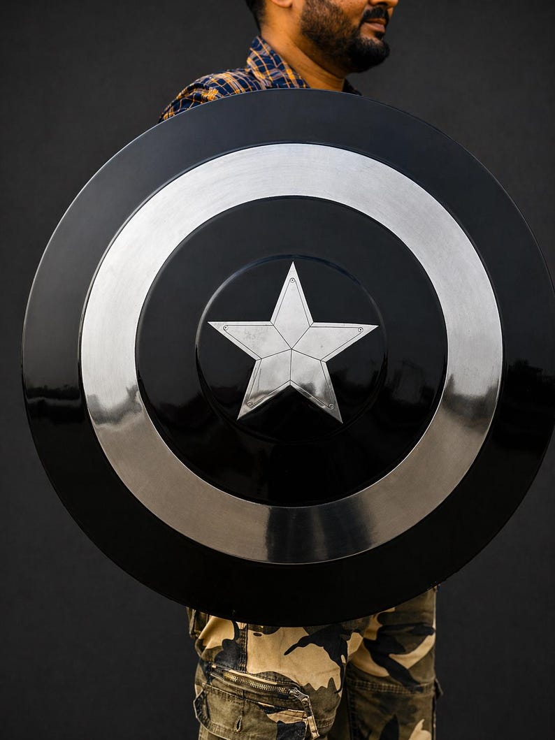 Puede incluir: Un escudo redondo negro y plateado, con una estrella plateada central. El escudo tiene un centro negro brillante, un anillo plateado y un borde exterior plateado. El escudo parece ser de metal.