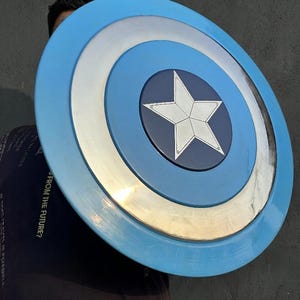 May include: A circular shield in shades of blue, silver, and white. The design includes a central white star and concentric rings. The outer edge is light blue. The shield's diameter is approximately 76 cm.