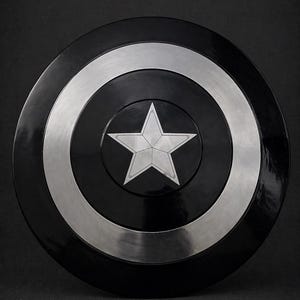 Puede incluir: Un escudo negro y plateado con una estrella blanca en el centro. El escudo tiene forma circular, con un anillo plateado alrededor de un centro negro. El borde exterior es negro y brillante. Un accesorio o art&iacute;culo de colecci&oacute;n.