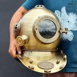 May include: A vintage brass diving helmet with the words "Anchor Engineering 1921" visible on the front. The helmet has a round viewing window and various brass fittings. The helmet is held by a person.