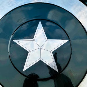 Puede incluir: Primer plano de un escudo met&aacute;lico con una gran estrella plateada de cinco puntas en el centro. La estrella est&aacute; sobre un fondo circular negro, rodeada por un borde plateado. El escudo tiene un acabado brillante.