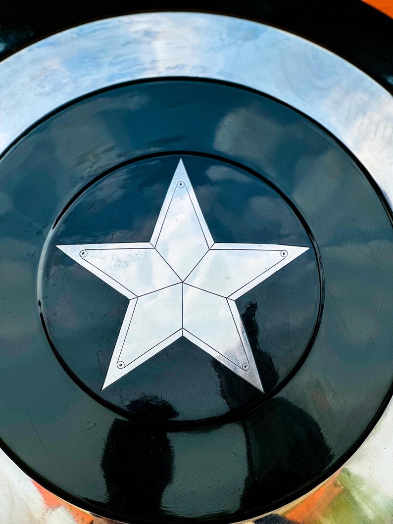 Puede incluir: Primer plano de un escudo met&aacute;lico con una gran estrella plateada de cinco puntas en el centro. La estrella est&aacute; sobre un fondo circular negro, rodeada por un borde plateado. El escudo tiene un acabado brillante.