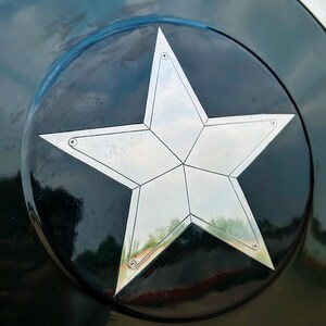 Puede incluir: Primer plano de un escudo negro y plateado con una gran estrella de cinco puntas en el centro. La estrella es plateada y tiene un dise&ntilde;o geom&eacute;trico. El escudo tiene un acabado brillante.