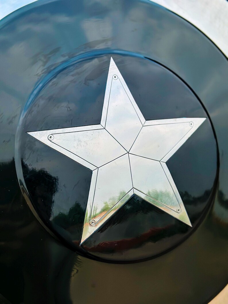 Puede incluir: Primer plano de un escudo negro y plateado con una gran estrella de cinco puntas en el centro. La estrella es plateada y tiene un dise&ntilde;o geom&eacute;trico. El escudo tiene un acabado brillante.