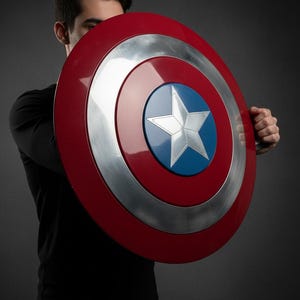 May include: A red, silver, and blue shield with a white star in the center. The shield is round with a red outer ring, a silver middle ring, and a blue center. The shield is held in front of a person.