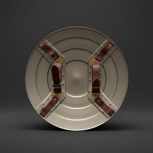 May include: A silver-colored, round shield with brown leather straps and brass-colored metal accents. The shield has a series of concentric circles and rivets. The straps are attached to the back of the shield.