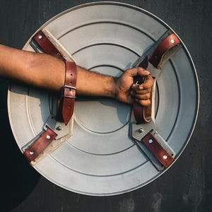 May include: A silver shield with brown leather straps and a handle. The shield has a circular design with concentric lines. The straps are riveted to the shield. The shield is held by a person's arm.