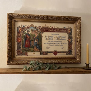 Peut inclure: Un certificat de mariage encadré avec des détails dorés ornés. Le certificat présente des illustrations d'un chevalier et d'une dame, avec le texte "Certificate of Marriage". Une bougie allumée est à droite. Le cadre mesure 50 cm de large.