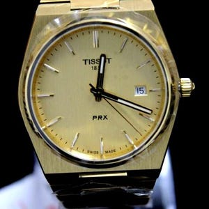 Puede incluir: Reloj Tissot PRX dorado con esfera dorada, ventana de fecha a las 3. Brazalete y bisel dorados. La esfera muestra "Tissot 1853 PRX" y "Swiss Made". Un accesorio elegante.