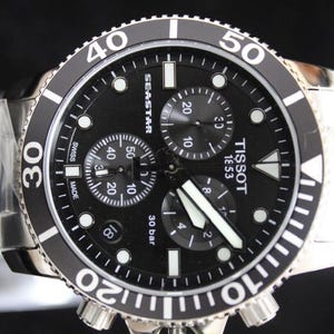 Op de afbeelding: Een Tissot Seastar horloge met een zwarte wijzerplaat en zilveren band. Het horloge heeft een zwarte draaibare bezel met witte markeringen, drie sub-wijzerplaten en lichtgevende wijzers. Waterdicht tot 30 bar.