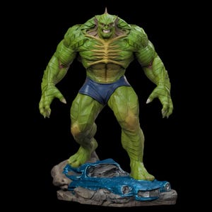 May include: A green, muscular figure with a menacing expression, wearing blue shorts, stands on a rock formation with a crushed blue car. The figure has a spiky head and is posed with arms outstretched. The statue is a collectible item.