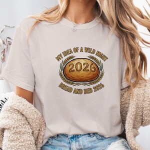 May include: A cream-colored t-shirt featuring the text "MY IDEA OF A WILD NIGHT" above an oval design with the number "2026" and the words "BREAD AND BED 2026". The design is surrounded by wheat stalks.