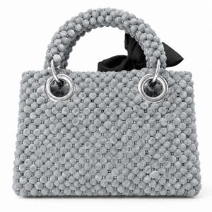 Puede incluir: Un bolso rectangular cubierto de pequeñas cuentas esféricas grises con textura de terciopelo. El bolso cuenta con un asa de cuentas a juego sujeta con grandes ojales metálicos plateados y un lazo de tela negra decorativo en un lateral.