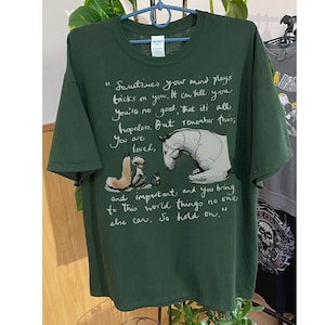 May include: Dark green t-shirt with a whimsical illustration of a horse and two figures, accompanied by handwritten text. The text reads "Sometimes your mind plays tricks on you...You are loved...So hold on."