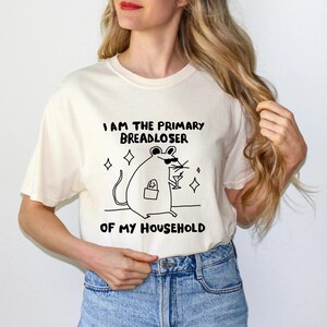 May include: Cream-colored t-shirt with a black graphic of a mouse wearing sunglasses and holding a cocktail. The text on the shirt reads "I AM THE PRIMARY BREADLOSER OF MY HOUSEHOLD."