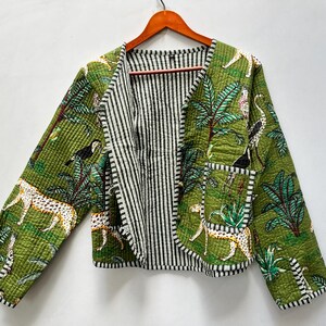 Puede incluir: Una chaqueta acolchada con fondo verde y un estampado de leopardos, aves tropicales y palmeras. La chaqueta tiene mangas largas, un bolsillo delantero y un forro de rayas blancas y negras visible en la parte delantera abierta y los bordes.