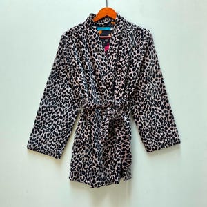 May include: A long-sleeved robe featuring a black and light pink leopard print pattern. The garment includes a matching fabric tie belt at the waist, a shawl collar, and a dark interior lining visible at the neckline, all displayed on a wooden hanger.