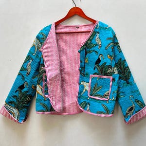 Puede incluir: Una chaqueta acolchada con base azul brillante y estampado de selva tropical que incluye tucanes, leopardos y palmeras. La chaqueta tiene cuello en V, mangas largas, un bolsillo frontal y un forro interior a rayas de color rosa y blanco.