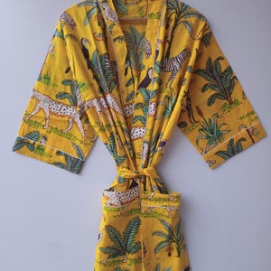 May include: A bright yellow robe featuring a repeating jungle print with leopards, zebras, toucans, and palm trees. The garment includes wide sleeves, a tie waist belt, and white piping trim along the front opening, cuffs, and pockets.