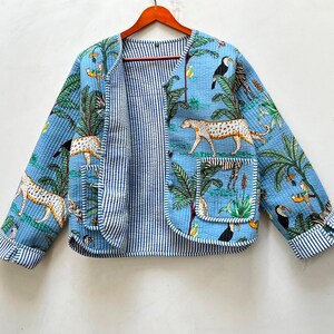 May include: A light blue quilted jacket featuring a jungle print with leopards, toucans, and palm trees. The jacket has a round neckline, long sleeves with striped cuffs, two front pockets, and a blue and white striped interior lining.