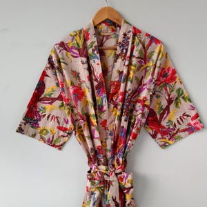 Könnte beinhalten: Ein hellbeiger Kimono-Bademantel mit einem lebhaften Blumen- und Vogelmuster in Rot-, Gelb-, Grün- und Violetttönen. Das Kleidungsstück hat weite Dreiviertelärmel, einen Schalkragen und einen passenden Bindegürtel aus Stoff in der Taille.