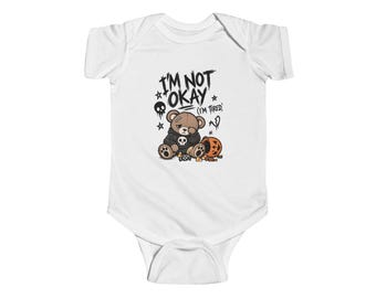 May include: A white short-sleeved bodysuit featuring a graphic of a brown teddy bear wearing a black hoodie with a skull print. The bear sits next to a pumpkin bucket and sweets. Text above reads I'M NOT OKAY (I'M TIRED) with stars and a skull.
