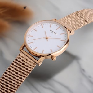 Puede incluir: Reloj de pulsera de oro rosa con esfera blanca y correa de malla. La esfera del reloj tiene marcadores y agujas de oro rosa, con el nombre de la marca "wyzer" impreso en la esfera. La correa es de diseño de malla fina.