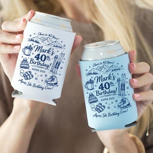 May include: Two beverage can coolers, one white and one light blue, are held in the image. Each cooler has a ski-themed design with the text "Mark's 40th Birthday!" and "Après Ski Birthday Crew!" The design includes skis, a mug, and snowflakes.
