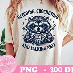 May include: Cream-colored t-shirt featuring a raccoon graphic holding yarn and knitting needles. The text above the raccoon reads "BITCHING, CROCHETING" and below reads "AND TALKING SHIT". The shirt also has the text "PNG - 300 DPI".