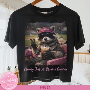 Può includere: T-shirt nero con un procione con occhiali da sole e una visiera "Game Day", che tiene in mano una bevanda e fa il segno della pace. Il testo "Literally Just A Bleacher Creature" è sotto l'immagine. La maglietta è appesa a una gruccia di legno.