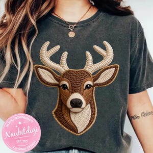 Puede incluir: Camiseta gris oscuro con un diseño de cabeza de ciervo bordada. El ciervo está representado en tonos marrones y beige, con costuras detalladas. La camiseta tiene un aspecto vintage y desgastado.