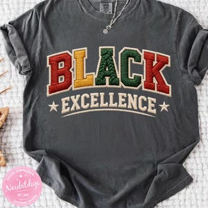 May include: Dark grey t-shirt featuring the embroidered words "BLACK EXCELLENCE". The word "BLACK" is in red, yellow, and green, and "EXCELLENCE" is in white with stars. A silver necklace is visible.