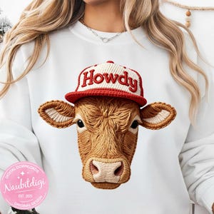 May include: White sweatshirt featuring an embroidered cow head wearing a red and white "Howdy" baseball cap. The cow's face is detailed with brown and tan thread, and the design is centered on the shirt.