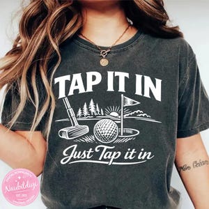 Puede incluir: Camiseta gris oscuro con un diseño blanco con temática de golf. El diseño incluye una pelota de golf, un palo, una bandera y las frases "TAP IT IN" y "Just Tap it in". La camiseta tiene un corte holgado y mangas cortas.