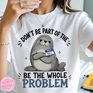 Puede incluir: Una camiseta blanca con un gráfico de una foca gris con los brazos cruzados, que lleva una etiqueta azul con el nombre "HELLO NEIL". El texto sobre la foca dice "DON'T BE PART OF THE" y el texto debajo dice "BE THE WHOLE PROBLEM".