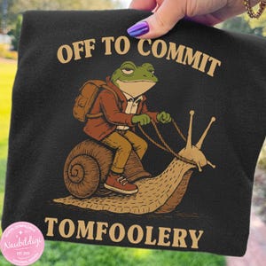 May include: Black t-shirt featuring a frog riding a snail, with the words "OFF TO COMMIT TOMFOOLERY". The frog is dressed in a jacket and backpack. A quirky and humorous design, perfect for those who enjoy a bit of fun.