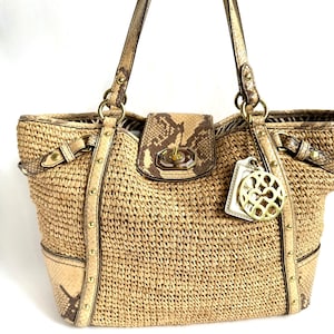 May include: A tan woven straw tote bag featuring snakeskin-patterned leather trim, gold-tone metal studs, and a central turn-lock closure. The bag includes two shoulder straps and a decorative gold-tone circular logo charm hanging from the side.
