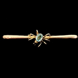 May include: A gold bar brooch featuring a central spider motif. The spider has a small, oval-shaped, light blue gemstone set into its body. The brooch has a polished metallic finish and a slender, horizontal design that extends outward from the spider.