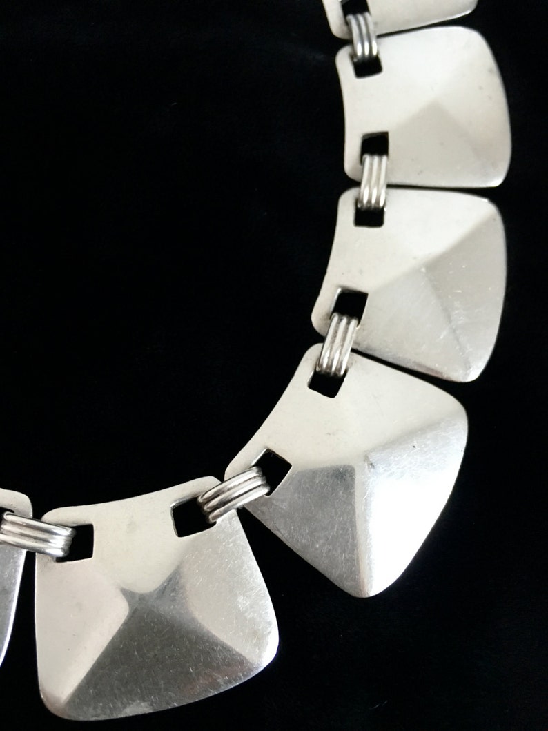 May include: A silver necklace with geometric, square-shaped links. The links are connected by a series of smaller, twisted silver rings.