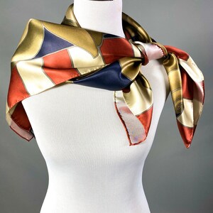 May include: A silk scarf with a geometric pattern in shades of gold, red, white, and blue. The scarf is tied around a white mannequin's neck.