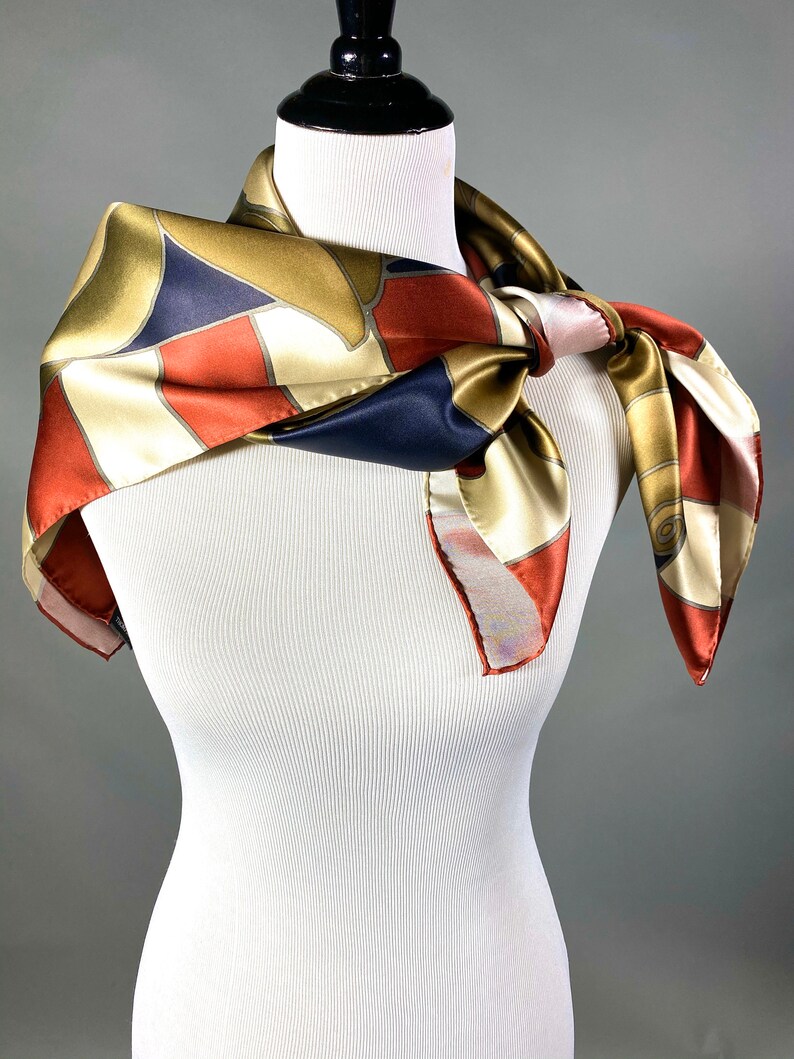 May include: A silk scarf with a geometric pattern in shades of gold, red, white, and blue. The scarf is tied around a white mannequin's neck.