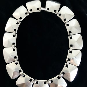 May include: A silver necklace with a geometric design. The necklace is made of a series of linked, square-shaped pieces with a raised, pyramid-like center.