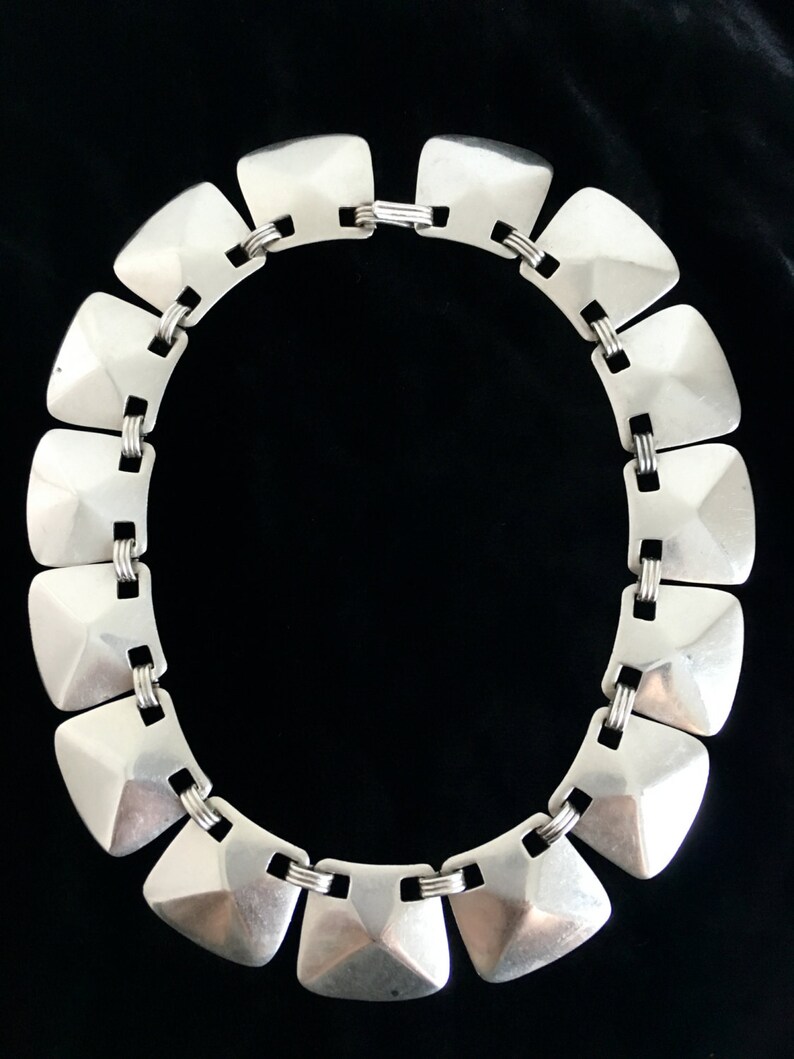 May include: A silver necklace with a geometric design. The necklace is made of a series of linked, square-shaped pieces with a raised, pyramid-like center.