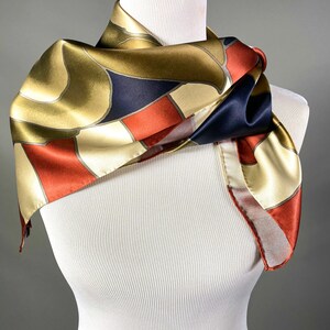 May include: A gold, red and navy blue patterned silk scarf. The scarf is folded and draped around a white mannequin.