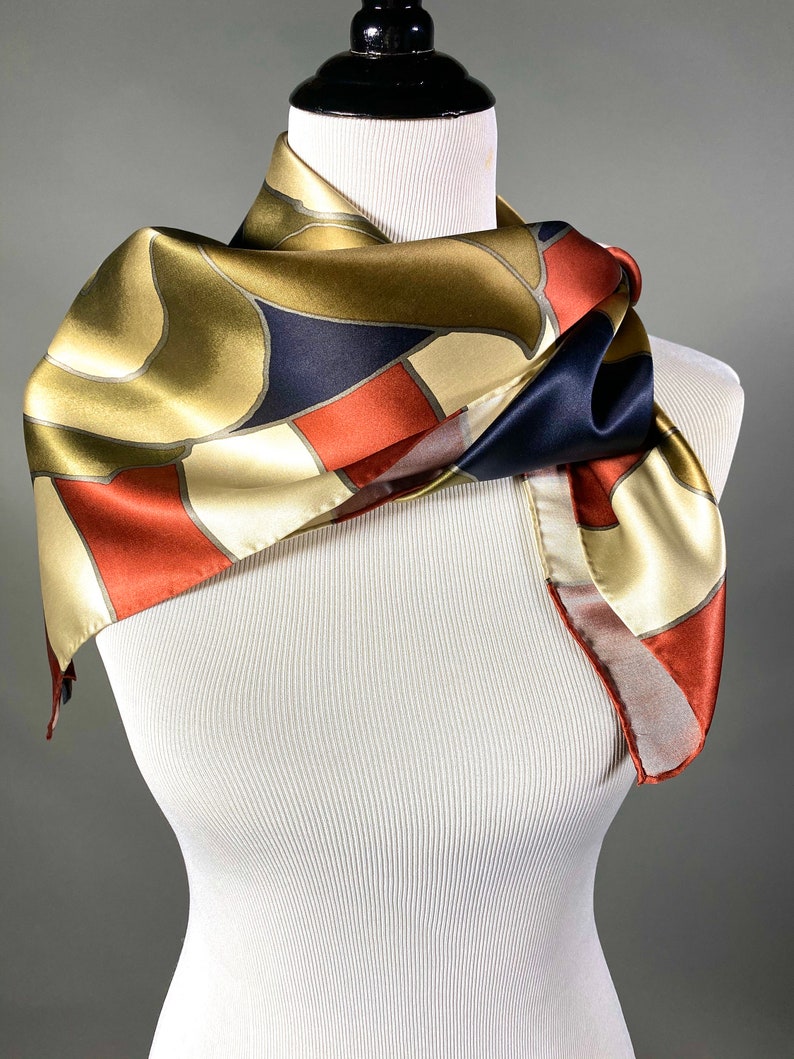 May include: A gold, red and navy blue patterned silk scarf. The scarf is folded and draped around a white mannequin.
