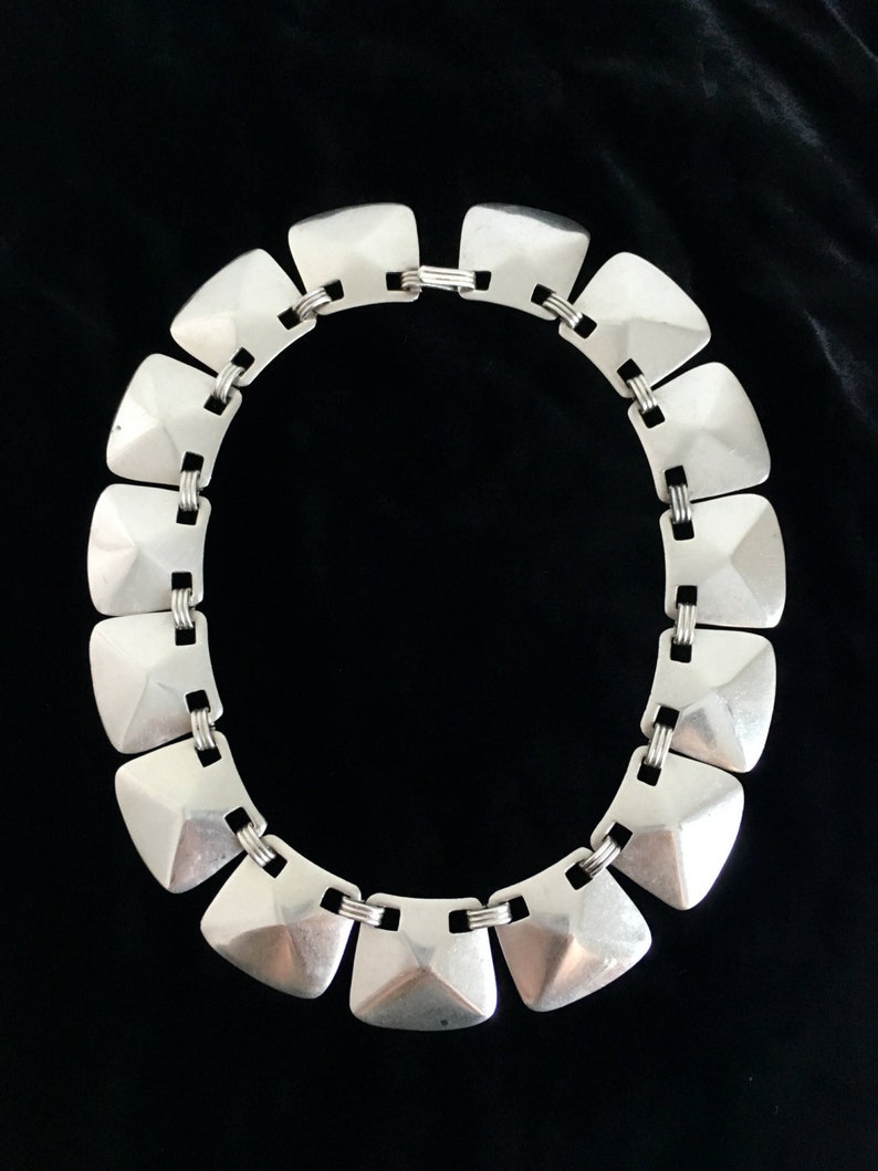 May include: A silver necklace with a geometric design. The necklace is made of a series of linked, square-shaped pieces with a raised, pyramid-like center.